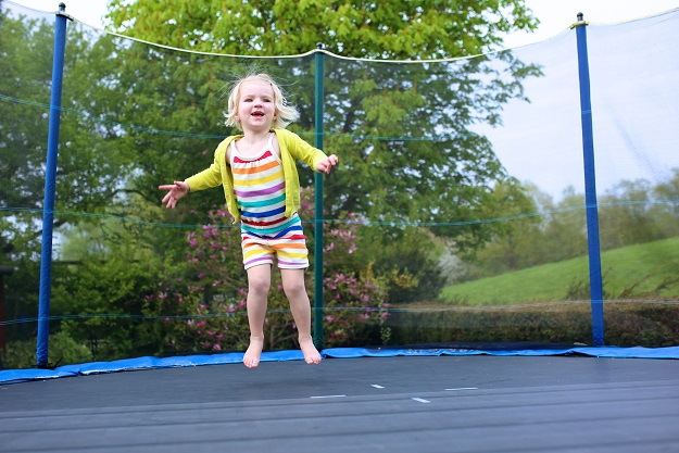 Happy little child, blonde curly toddler girl jumping on trampoline in the garden at the backyard of the house on a sunny summer day